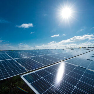 Solar panes in a solar park used for clean energy production. Solar panel under blue sky with sun. Green grass and cloudy sky. Alternative energy concept to reduce global warming and climate change